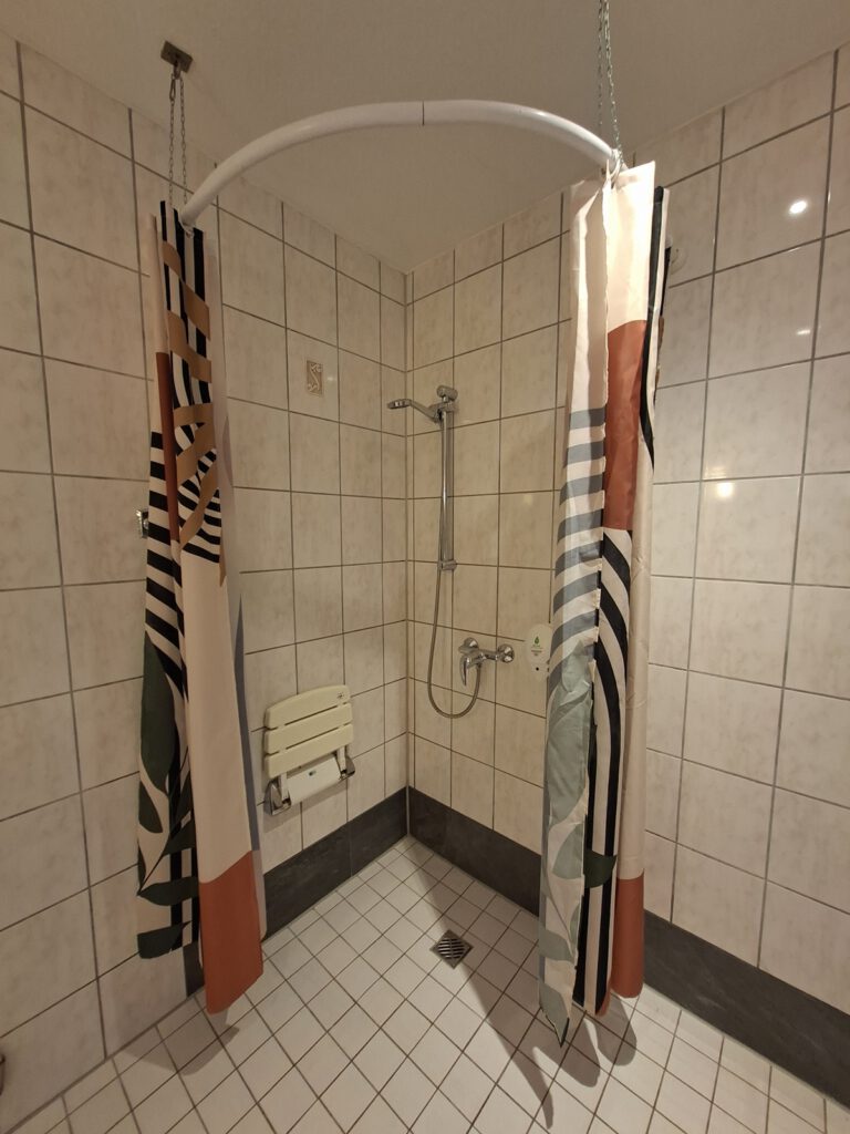 Hotel Sonne Karlsruhe. Eine barrierefreie, rollstuhlgeeignete Dusche mit ausklappbarer Sitzmöglichkeit und Seifenspender in Griffhöhe.